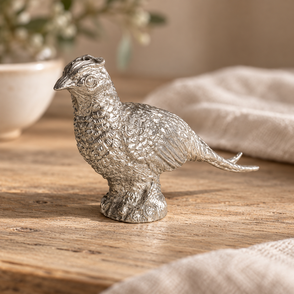 Pewter pheasant shaker on a pale wooden table beside a linen napkin