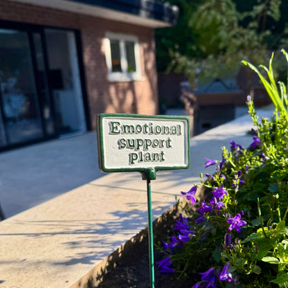 A small rectangular garden sign with the words 'Emotional support plant' in raised dark lettering on a light background, mounted on a green stake in a garden bed with purple flowers and a blurred house in the background.
