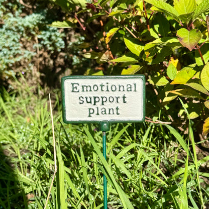A small rectangular garden sign with a white face and green border that reads 'Emotional support plant,' mounted on a green stake in a grassy garden with leafy plants in the background.