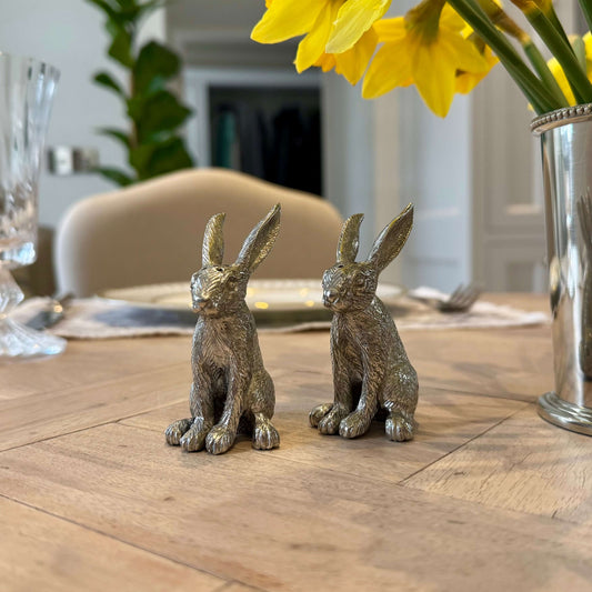 Handcrafted pewter rabbit salt and pepper shakers on a wooden dining table with a reflective vase of yellow daffodils; luxury tableware by Woollaston White