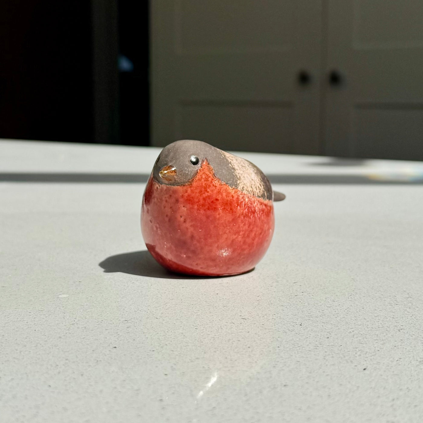 A small bird figurine with a glossy red body and gray head, sitting on a light gray tabletop in natural sunlight.