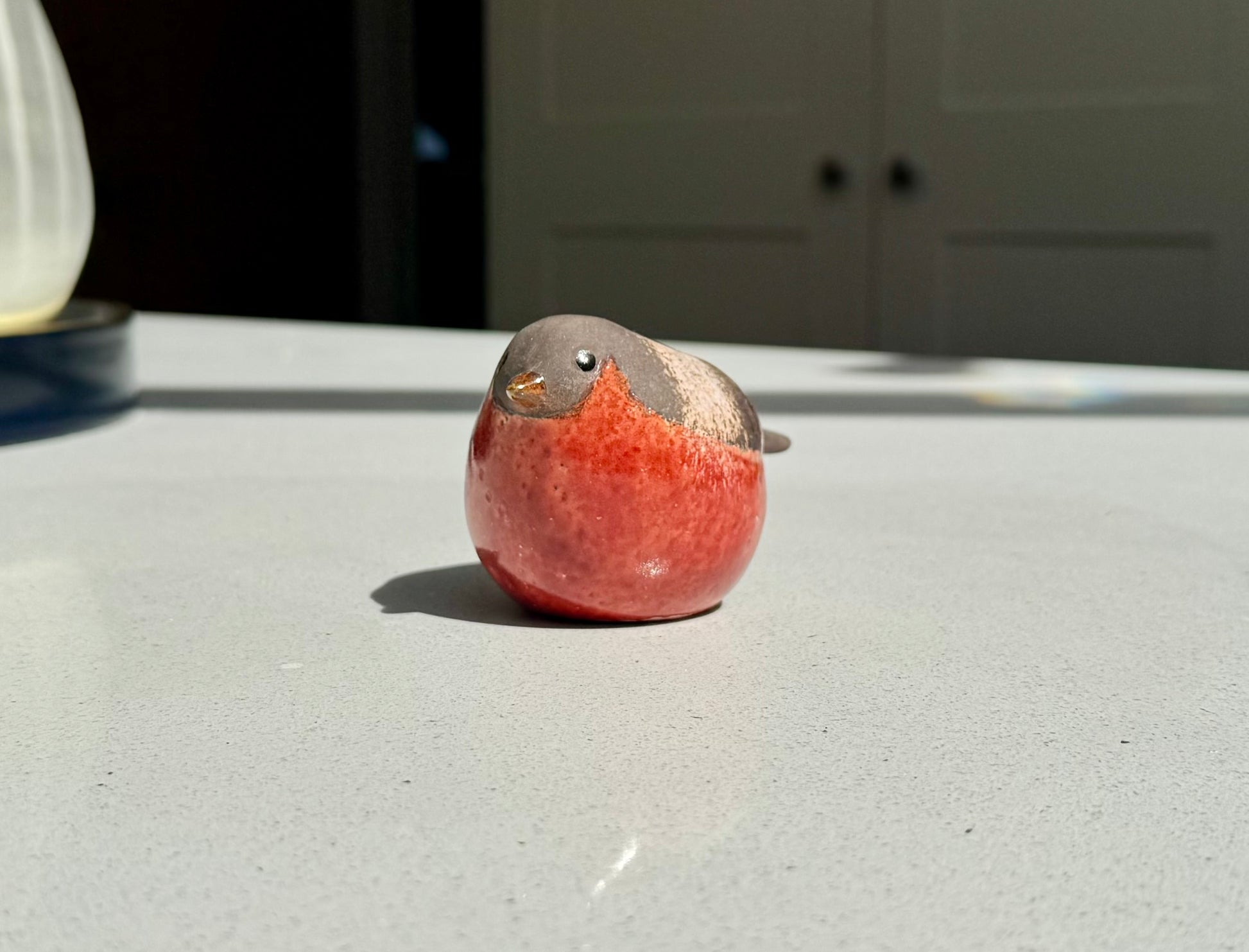A small bird figurine with a glossy red body and gray head, sitting on a light gray tabletop in natural sunlight.