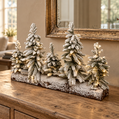 A decorative tabletop arrangement of five frosted Christmas trees on a rustic wooden console table in a warm living room setting.