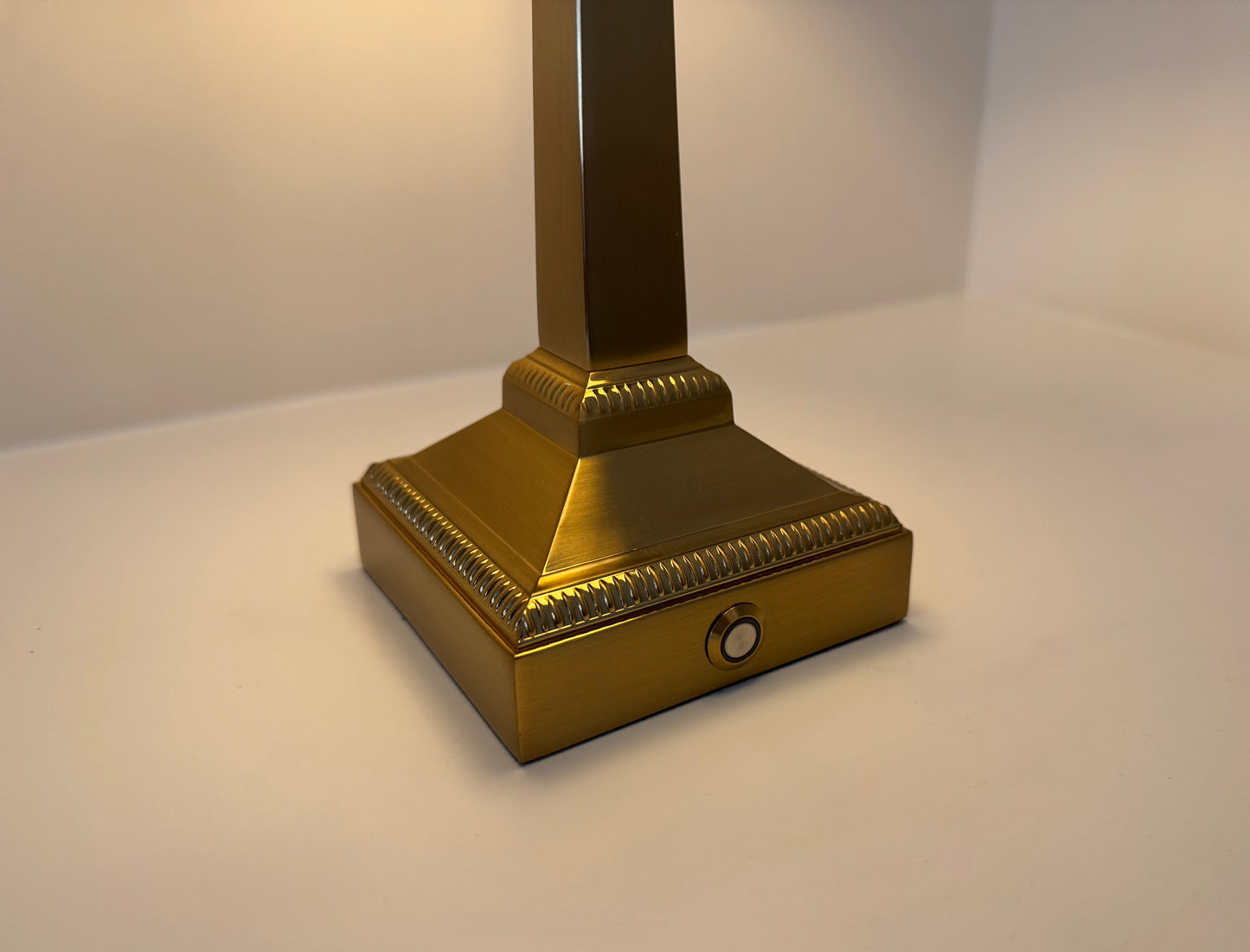 A close-up of a decorative brass lamp base with a square pedestal design and a vertical stem, set against a plain light background.