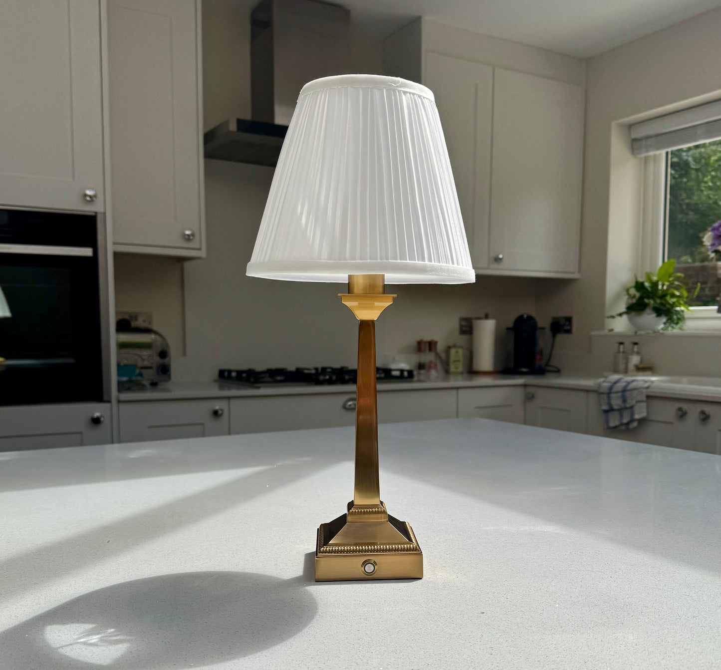 A tall rechargeable table lamp with a white pleated fabric shade and a polished brass base, set on a white kitchen countertop.